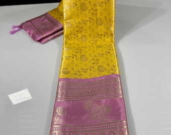 May include: A vibrant yellow saree with intricate floral patterns and a contrasting pink border. The saree is folded, showcasing its detailed design and texture. The pink border features gold-colored floral motifs and tassels.