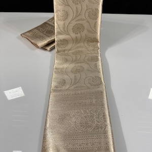 May include: A folded, beige-colored textile with intricate floral and paisley patterns. The fabric has a subtle sheen, and the design includes detailed borders. The textile is likely a decorative fabric or a piece of clothing.