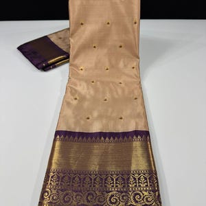 May include: A beige silk saree with gold embroidery and a purple border. The saree features small gold floral accents throughout the body and a detailed gold and purple patterned border. A matching folded piece of fabric is visible.