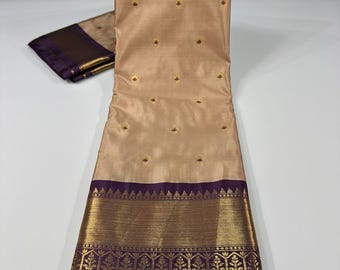 May include: A beige silk saree with gold embroidery and a purple border. The saree features small gold floral accents throughout the body and a detailed gold and purple patterned border. A matching folded piece of fabric is visible.
