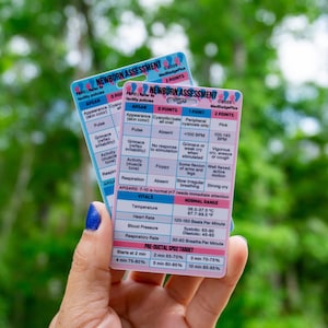 May include: Two newborn assessment cards with pink and blue backgrounds are held up. The cards list APGAR scores, vitals, and pre-ductal SpO2 targets. The text on the cards is in English, with measurements in Fahrenheit and beats per minute.