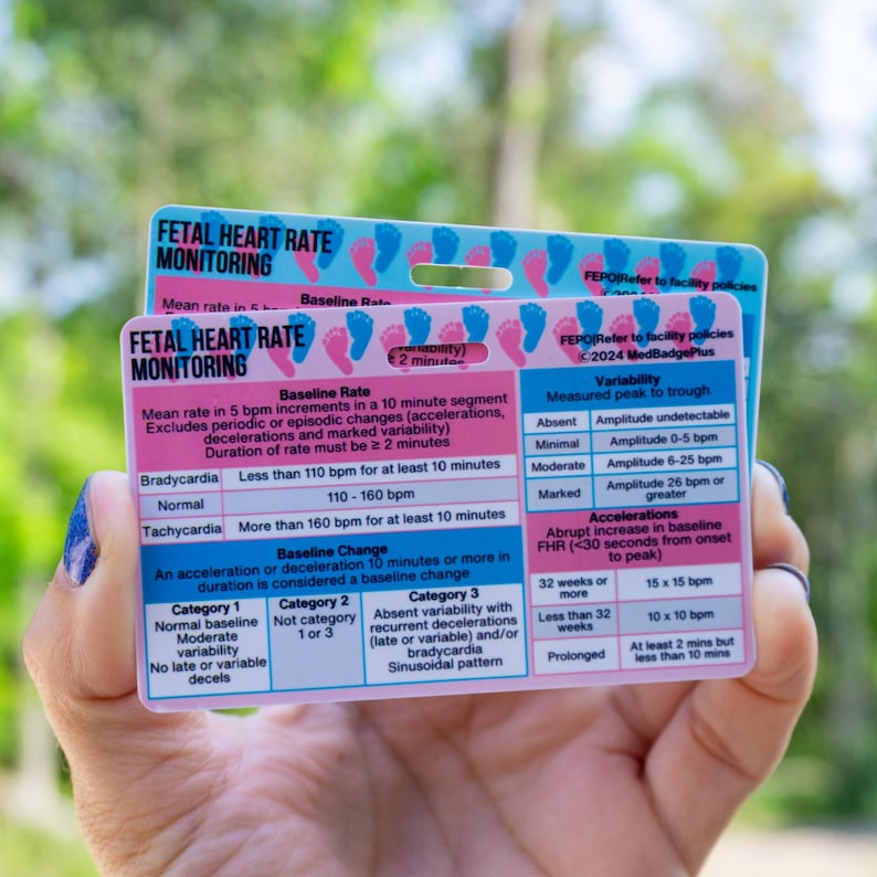 May include: Two rectangular cards with medical information about fetal heart rate monitoring. The cards are white, pink, and blue with text and diagrams. One card is held in a hand with blue nail polish. The cards include information on baseline rate, variability, and accelerations.