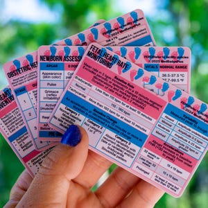 May include: A hand holds several pink and white cards with medical information. The cards have text about fetal heart rate monitoring, newborn assessment, and obstetric hemorrhage. The cards have blue and pink footprints.
