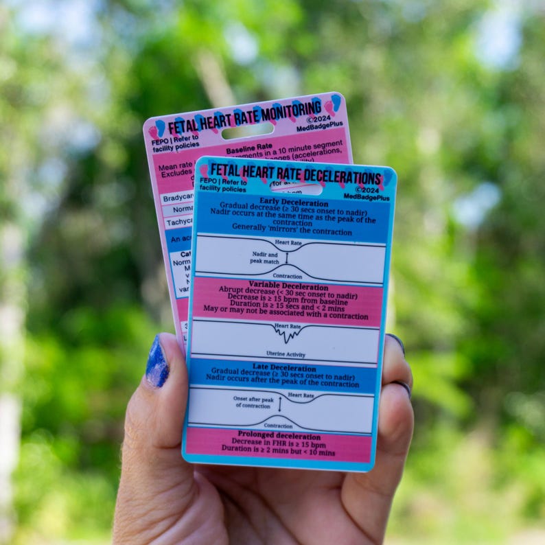 May include: Two rectangular cards with medical information are held in front of a blurred green background. The cards are pink and blue, with text and diagrams about fetal heart rate monitoring and decelerations. The cards are labeled "FETAL HEART RATE MONITORING" and "FETAL HEART RATE DECELERATIONS".