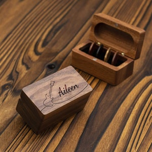 Könnte beinhalten: Zwei Holzkisten, eine mit einer Gitarre und dem Namen "Aileen" auf dem Deckel graviert, die andere mit einem Klappdeckel und einem Fach für Gitarrenplektren.