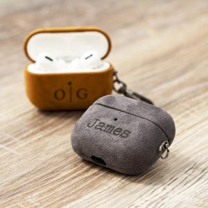 May include: Two personalized AirPod cases. One is brown with the initials "o|g" engraved, and the other is gray with the name "James" engraved. Both cases are made of a soft, suede-like material and are open, revealing the white AirPods inside.