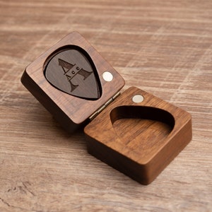 K&ouml;nnte beinhalten: Eine Holzkiste mit einem Scharnierdeckel, die f&uuml;r ein Gitarrenplektrum gedacht ist. Die Kiste ist aus dunklem Holz und hat eine nat&uuml;rliche Oberfl&auml;che. Der Deckel ist ge&ouml;ffnet und zeigt ein Gitarrenplektrum mit dem eingravierten Buchstaben "A". Das Plektrum ist aus dunklem Holz und hat eine nat&uuml;rliche Oberfl&auml;che.