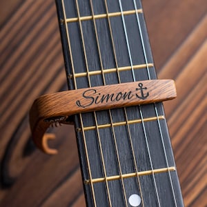 K&ouml;nnte beinhalten: Ein h&ouml;lzerner Gitarrenkapodaster mit dem Namen "Simon" und einem eingravierten Anker-Symbol. Der Kapodaster ist am Hals einer Gitarre befestigt.