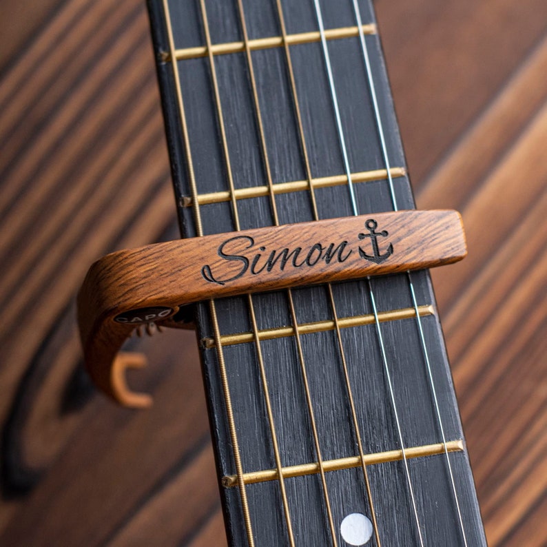 K&ouml;nnte beinhalten: Ein h&ouml;lzerner Gitarrenkapodaster mit dem Namen "Simon" und einem eingravierten Anker-Symbol. Der Kapodaster ist am Hals einer Gitarre befestigt.