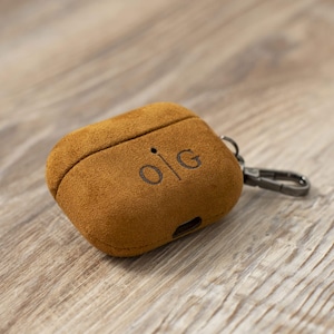 Könnte beinhalten: Eine braune Wildleder-AirPod-Hülle mit den Initialen "OIG" auf der Vorderseite. Die Hülle hat einen Metallverschluss und liegt auf einer Holzoberfläche. Die Hülle dient zum Schutz und Transport von kabellosen Ohrhörern.