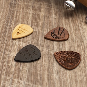 K&ouml;nnte beinhalten: Vier Holz-Picks mit verschiedenen Designs. Ein Pick ist schwarz mit dem Text "Elly loves you". Ein Pick ist braun mit einem Fingerabdruck-Design. Ein Pick ist braun mit einem Schlagzeug- und Schlagzeugst&ouml;cken-Design. Ein Pick ist hellbraun mit dem Text "I will always pick you" und einem kleinen Herzen.