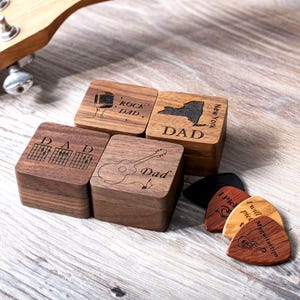 May include: Four square wooden boxes with engraved designs including a microphone, New York state map, guitar chords, and a guitar, labeled with the word Dad. Three guitar picks are placed nearby, featuring text and a small guitar illustration.