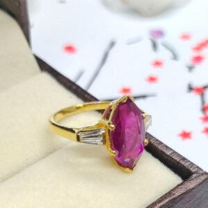 Könnte beinhalten: Ein goldener Ring mit einem großen, marquiseförmigen, magentafarbenen Edelstein als Mittelpunkt. Der Ring hat ein goldenes Band und wird von zwei kleineren, rechteckigen, klaren Edelsteinen flankiert. Der Ring wird in einer braunen Schmuckschatulle präsentiert.