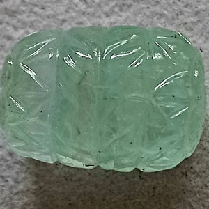 May include: A carved green gemstone with a detailed floral pattern. The gemstone is smooth and polished, with a translucent appearance.