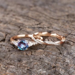 Könnte beinhalten: Ein Verlobungsring-Set aus Roségold mit einem runden, teal-farbenen Mittelstein und zwei kleineren weißen Steinen auf jeder Seite. Das passende Band hat ein verdrehtes Design mit kleinen, klaren Steinen. Text: ASGemsNjewelry.