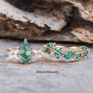 Könnte beinhalten: Zwei goldene Ringe. Ein Ring hat einen großen, rautenförmigen grünen Edelstein mit weißen Akzentsteinen. Der andere Ring hat ein zartes Design aus grünen, blattförmigen Edelsteinen. Die Ringe sind vor einem neutralen Hintergrund platziert.