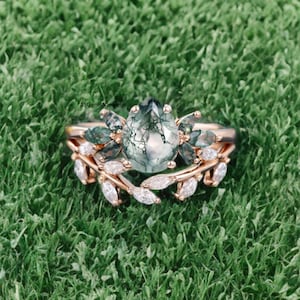 Könnte beinhalten: Ein roségoldenes Ring-Set mit einem birnenförmigen Moosachat-Mittelstein und einem blattförmigen Band mit weißen Akzentsteinen. Der Ring liegt auf grünem Gras.