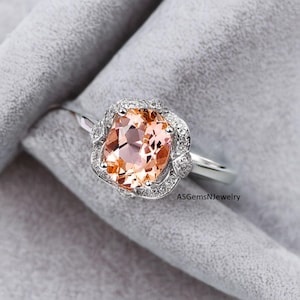 Può includere: Un anello d'argento con una grande pietra preziosa ovale color pesca, circondata da un alone di pietre più piccole e trasparenti. L'anello è posto su uno sfondo grigio. Il testo "ASGemsNjewelry" è visibile.