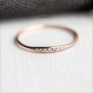 May include: A delicate rose gold ring with a curved design. The ring is adorned with a series of small, sparkling diamonds set along the curve. The ring is set on a white surface.
