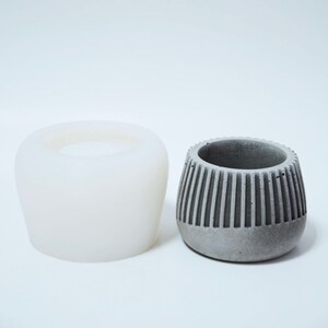 May include: A white silicone mold and a gray concrete planter with a ribbed design. The planter is slightly larger than the mold.