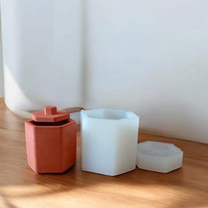 May include: An orange hexagonal container with a lid, a white hexagonal mould, and a small white hexagonal piece sit on a wooden surface. The objects are likely for crafting or storage.