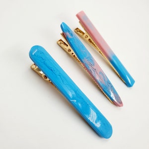 Op de afbeelding: Drie haarclips met gouden metalen sluitingen. De clips zijn gemaakt van hars en hebben een marmermotief. E&eacute;n clip is blauw, &eacute;&eacute;n is roze en &eacute;&eacute;n is een mix van roze en blauw.
