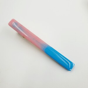 Op de afbeelding: Een lange, dunne, blauwe en roze haarspeld. De speld is gemaakt van plastic en heeft een kleurverloop van roze naar blauw.