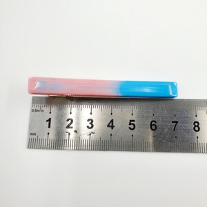 Op de afbeelding: Een roze en blauwe plastic haarspeld met een lengte van ongeveer 5 centimeter. De speld is geplaatst op een liniaal ter referentie van de grootte.