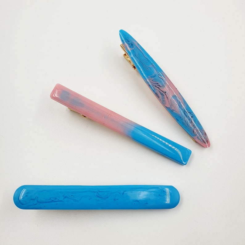 Op de afbeelding: Drie haarclips in verschillende stijlen. E&eacute;n is een lange, dunne, blauwe en roze marmeren clip. Een andere is een kortere, dikkere, blauwe en roze ombre clip. De laatste is een effen blauwe clip.