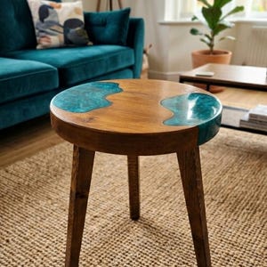 Può includere: Un tavolino rotondo in legno con tre gambe affusolate e una superficie con venature naturali. Due sezioni distinte e irregolari di resina traslucida color verde acqua sono incastonate nel piano del tavolo, creando una finitura liscia e uniforme.