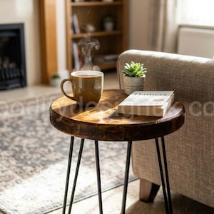 May include: A round wooden side table with a dark brown stained finish and three thin black metal hairpin legs. On the table sits a steaming tan ceramic mug and a closed book with a white cover. The table surface shows a visible wood grain.