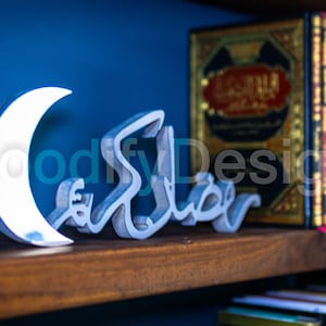 May include: A silver crescent moon and the Arabic word "Ramadan" in silver, displayed on a wooden shelf with books in the background.