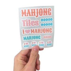 K&ouml;nnte beinhalten: Ein Bogen Aufkleber mit den W&ouml;rtern "MAHJONG", "Tiles" und "I &hearts; MAHJONG" in verschiedenen Schriftarten und Farben. Zus&auml;tzliche Aufkleber sagen "LUCKY" und "MAHJONG BAM!". Die Aufkleber werden von einer Hand vor wei&szlig;em Hintergrund gehalten.