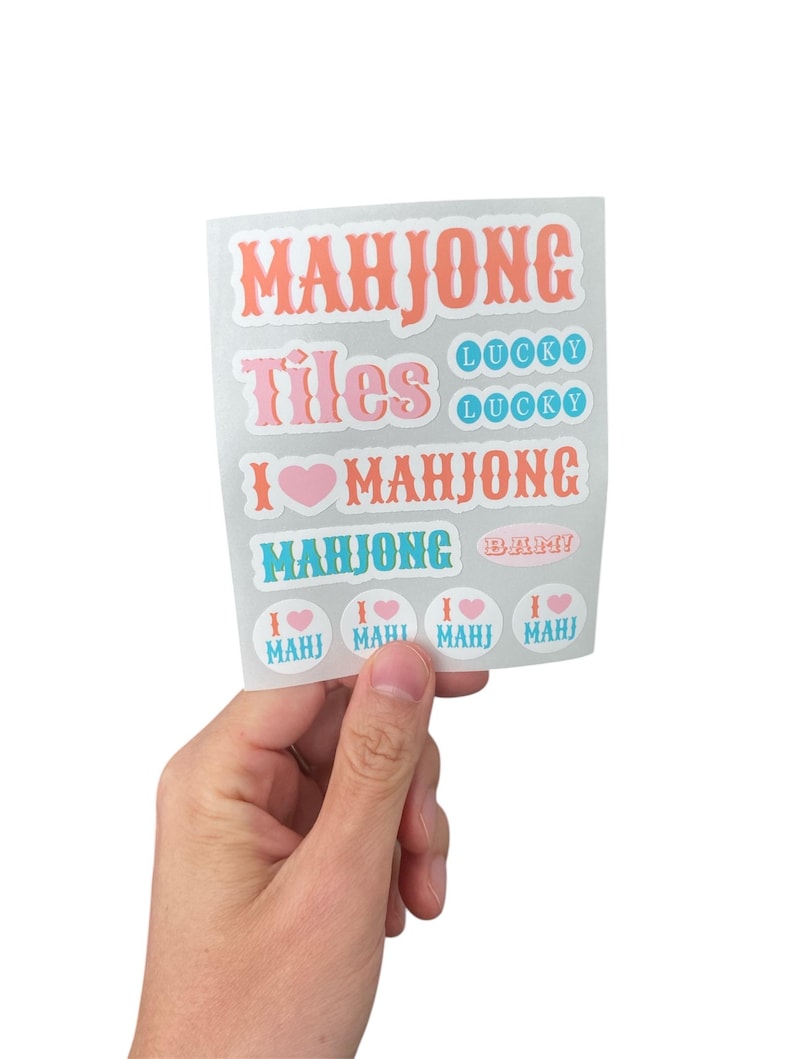 K&ouml;nnte beinhalten: Ein Bogen Aufkleber mit den W&ouml;rtern "MAHJONG", "Tiles" und "I &hearts; MAHJONG" in verschiedenen Schriftarten und Farben. Zus&auml;tzliche Aufkleber sagen "LUCKY" und "MAHJONG BAM!". Die Aufkleber werden von einer Hand vor wei&szlig;em Hintergrund gehalten.