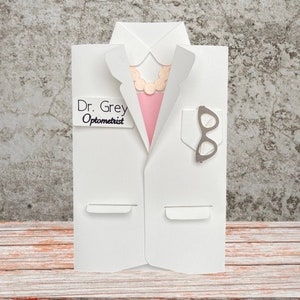 May include: A white paper card shaped like a doctor's coat, with a pink shirt and a necklace peeking out. It has a pocket with glasses and a name tag that reads "Dr. Grey Optometrist". The card is on a wooden surface.