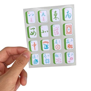 K&ouml;nnte beinhalten: Ein Blatt mit Mahjong-Fliesenaufklebern, das von einer Hand gehalten wird. Die Aufkleber sind rechteckig mit einem hellgr&uuml;nen Rand und zeigen verschiedene Symbole und Zahlen in mehreren Farben, darunter Blau, Rosa und Orange.