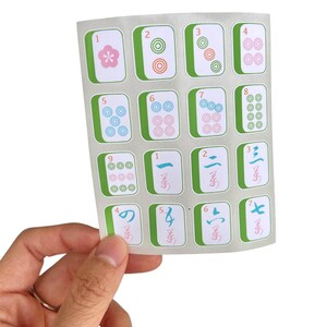 K&ouml;nnte beinhalten: Ein Bogen mit Mahjong-Fliesenaufklebern. Jeder Aufkleber zeigt eine Zahl oder ein Symbol mit einem gr&uuml;nen Rand und wei&szlig;em Hintergrund. Die Kacheln werden von einer Hand vor einem wei&szlig;en Hintergrund gehalten.