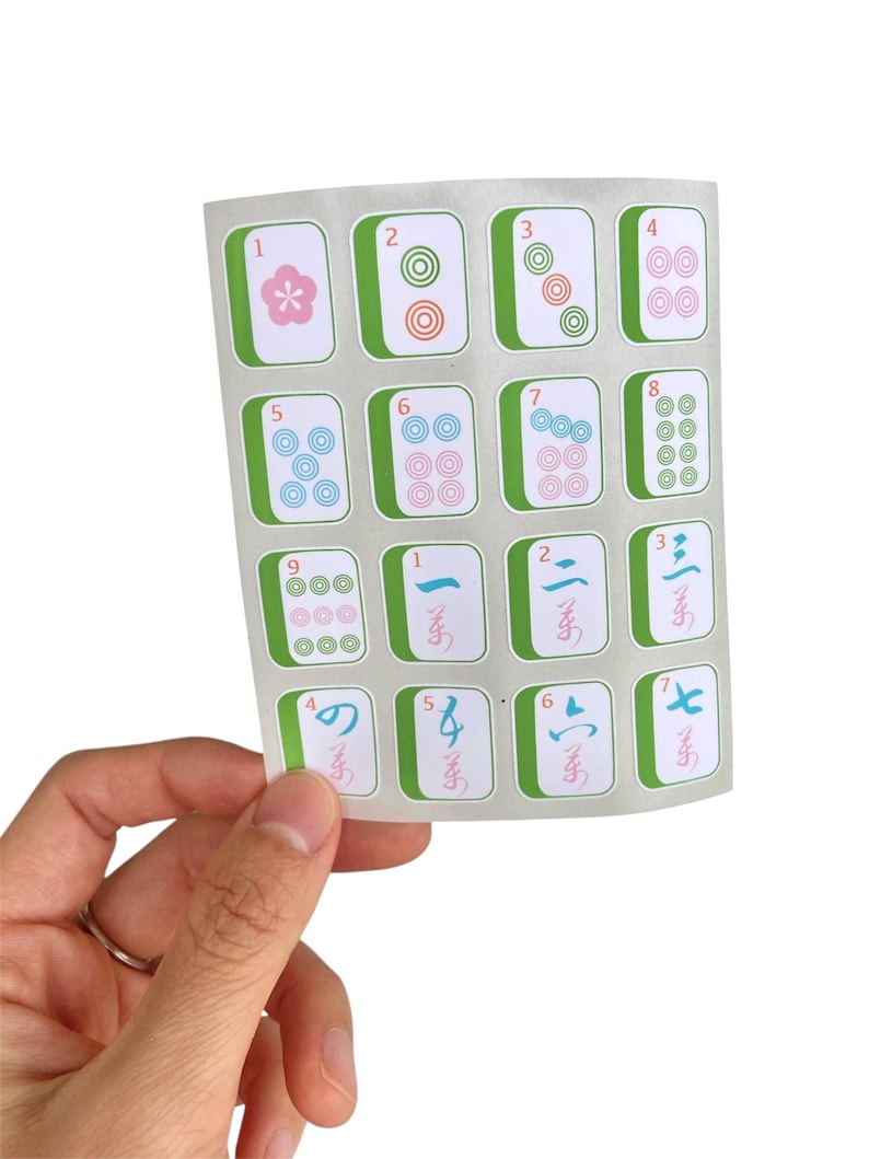 K&ouml;nnte beinhalten: Ein Bogen mit Mahjong-Fliesenaufklebern. Jeder Aufkleber zeigt eine Zahl oder ein Symbol mit einem gr&uuml;nen Rand und wei&szlig;em Hintergrund. Die Kacheln werden von einer Hand vor einem wei&szlig;en Hintergrund gehalten.