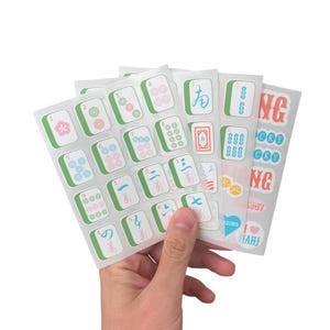 K&ouml;nnte beinhalten: Eine Hand h&auml;lt mehrere Stickerb&ouml;gen mit Mahjong-Kachel-Designs. Die Aufkleber haben einen wei&szlig;en Hintergrund mit gr&uuml;nen, rosa und blauen Akzenten. Einige Aufkleber tragen die Aufschrift "JONG" und "CKY".