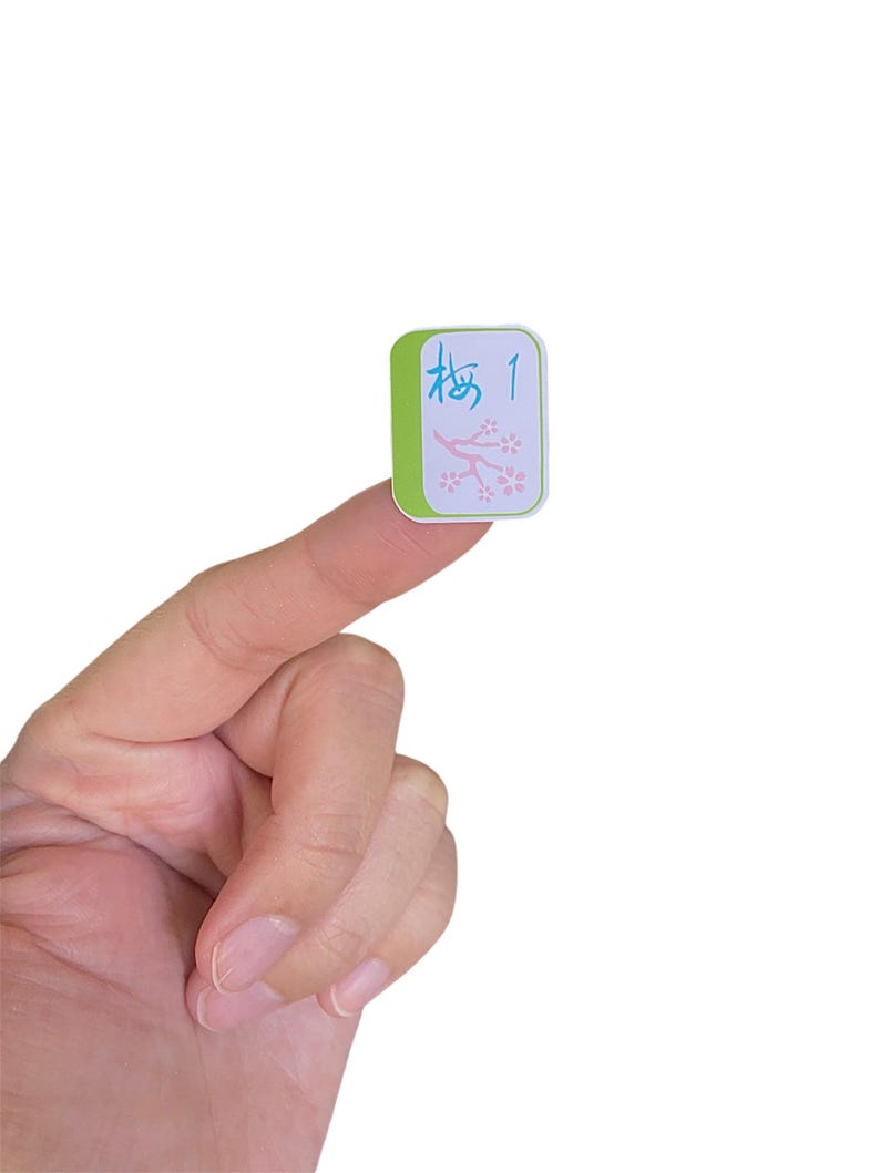 K&ouml;nnte beinhalten: Eine gr&uuml;ne und wei&szlig;e Mahjong-Kachel mit der Zahl 1 und japanischen Schriftzeichen. Die Kachel hat einen hellblauen Rand und ein rosa Blumenmuster. Die Kachel wird zwischen zwei Fingern gehalten.