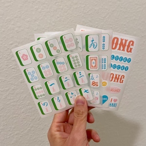 K&ouml;nnte beinhalten: Eine Hand h&auml;lt mehrere Stickerb&ouml;gen mit Mahjong-Steinen und Text. Die Aufkleber haben einen wei&szlig;en Hintergrund mit gr&uuml;nen, blauen, roten und rosa Akzenten. Einige Aufkleber sagen "ONG" und "LUCKY".