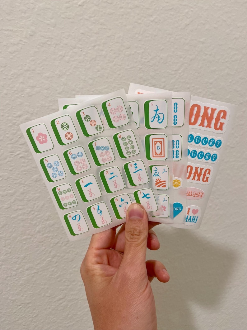 K&ouml;nnte beinhalten: Eine Hand h&auml;lt mehrere Stickerb&ouml;gen mit Mahjong-Steinen und Text. Die Aufkleber haben einen wei&szlig;en Hintergrund mit gr&uuml;nen, blauen, roten und rosa Akzenten. Einige Aufkleber sagen "ONG" und "LUCKY".