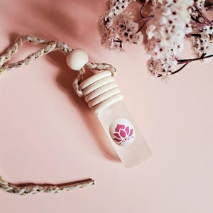 Può includere: Una bottiglia di vetro bianco con un design a forma di fiore di loto rosa, appesa a una corda beige con un perno di legno. La bottiglia è su uno sfondo rosa.