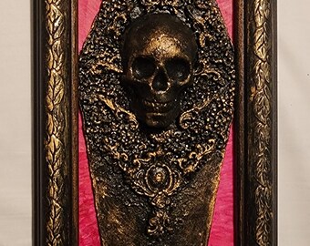May include: A framed, gothic-style wall art piece. The artwork features a detailed, gold-colored skull set against a textured, dark background, framed within a black and gold ornate border. The background is a vibrant pink.
