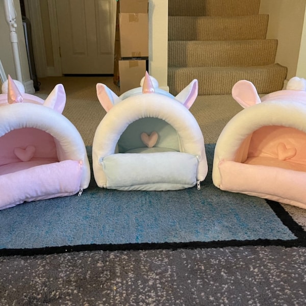 Könnte beinhalten: Drei tierförmige Betten in Form eines Einhorns in rosa, blau und weiß. Jedes Bett hat ein herzförmiges Kissen im Inneren und einen Reißverschluss.
