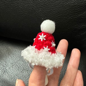 May include: A red crocheted Santa hat with a white pom pom and snowflake accents. The hat is worn on a finger as a ring.