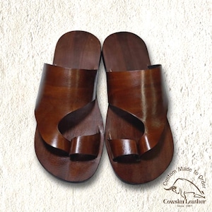 May include: A pair of dark brown leather slide sandals featuring a wide strap across the top and a separate loop for the big toe. The sandals have a smooth, polished finish and a flat sole. A logo with a bull and the text Custom Made to Order Cowshu Leather Since 1997 is visible in the bottom right corner.