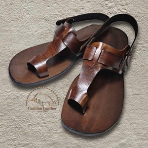 May include: A pair of dark brown leather sandals featuring a toe loop design, wide straps across the midfoot, and an adjustable ankle strap with a metal buckle. The logo for Cowshu Leather is visible on the left sandal, featuring a bull icon.
