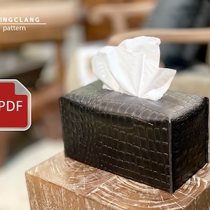 May include: A black, rectangular tissue box cover with a crocodile skin effect, dispensing white tissues. The cover is on a wooden surface. The text "CLINGCLANG pattern" and a PDF icon are in the upper left corner.