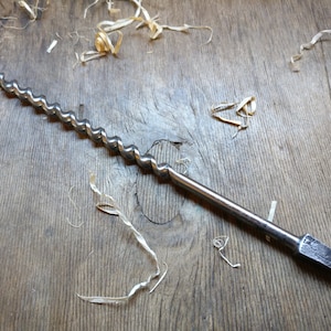 May include: A silver metal drill bit with a long, twisted shaft and a pointed tip. The drill bit is lying on a wooden surface with wood shavings scattered around it.